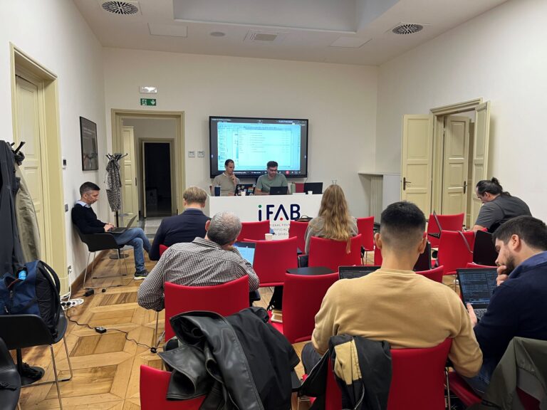 People sitting in a conference room at iFAB, a presentation in progress with two speakers seated at the front desk. The audience members are seated on red chairs, some working on laptops, while others are listening to the presentation. The room has light-colored walls, wooden flooring, and an organized setup with coats and bags placed to the side.